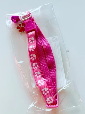 Rosa katt-halsband med tassmönster - Säljer ett rosa katt-halsband i nylon med vita tassavtryck och ett litet rosa klockhänge. Halsbandet har ett metallspänne och en söt tassformad bricka. Perfekt accessoar för din katt!