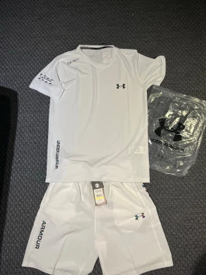 Vitt träningsset från Under Armour - Snyggt vitt träningsset från Under Armour med t-shirt och shorts. Perfekt som träningskläder men också till sommaren. Storleken är L men den passar perfekt som M. Ifall ni har några funderingar så är det bara att fråga.