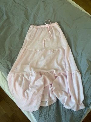 Ljusrosa volangkjol med snörning - Söt ljusrosa kjol med volang och snörning i midjan. Kjolen har en luftig och flowy passform, perfekt för varma dagar. Tillverkad i ett mjukt tyg som känns skönt mot huden. Passar dig som gillar en feminin och avslappnad stil.