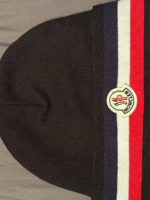 Moncler mössa - Snygg svart mössa från Moncler med klassiska ränder i blått, vitt och rött samt broderad logotyp framtill. Stickad modell i mjukt material som sitter skönt på huvudet. Perfekt accessoar för att lyfta din stil.