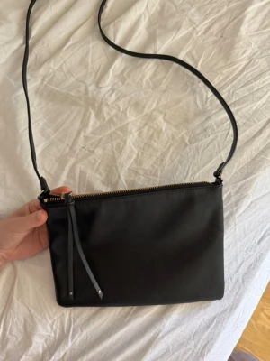 Svart axelväska - En stilren svart axelväska i slätt material med guldfärgad dragkedja och smal axelrem. Väskan har enkel design och en dekorativ dragkedjetofs. Perfekt för dig som gillar en clean och modern look.