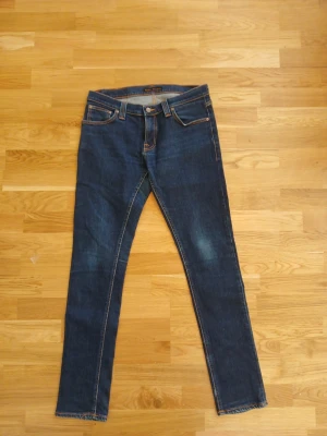 Mörkblå slim fit jeans från Nudie - Snygga mörkblå slim fit jeans från Nudie med klassiska fem fickor och orangea kontrastsömmar. Jeansen har en rak passform och är tillverkade i denim med lätt slitning på låren. Perfekta för en stilren och avslappnad look. Pris kan diskuteras