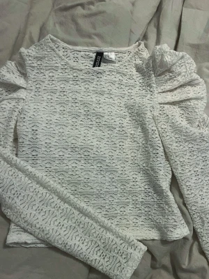 Vit spetsblus med puffärm från Divided - Superfin vit blus från Divided med lång ärm och puffiga axlar. Blusen är helt täckt av ett vackert spetsmönster och har en rund halsringning. Perfekt att styla med jeans eller kjol för en trendig look.