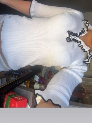 Vit ribbad topp med volangkant - Trendig vit ribbad topp med svarta volangdetaljer längs halsringning och ärmslut. Toppen har lång ärm och en figurnära passform som ger en snygg siluett. Perfekt för dig som gillar en stilren men ändå lekfull look.