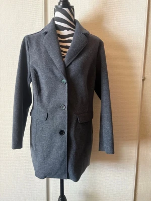 Mörkgrå klassisk ullrock - Stilren mörkgrå rock med tre knappar framtill och klassisk krage. Rocken har två fickor med lock och är tillverkad i ett ull-liknande material. Passar perfekt för en snygg och tidlös look under kallare dagar.