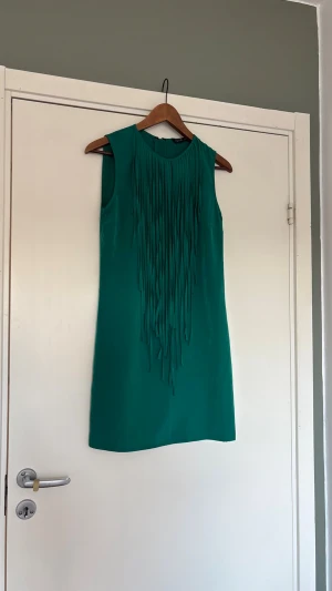 Turkos  ärmlös klänning med fransar - Snygg turkos ärmlös klänning med långa fransdetaljer framtill. Klänningen har en rak, kort passform och rund halsringning. 