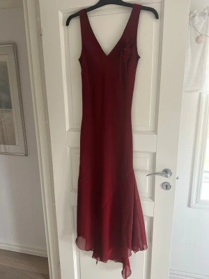 Vinröd långklänning med blomdetalj - Elegant vinröd långklänning med v-ringning fram och back och en dekorativ blomma. Klänningen har ett lätt, flytande tyg med svart undertyg/underklänning och asymmetrisk nederkant som ger en snygg siluett. Perfekt för speciella tillfällen tex bal. Endast använd en gång. 