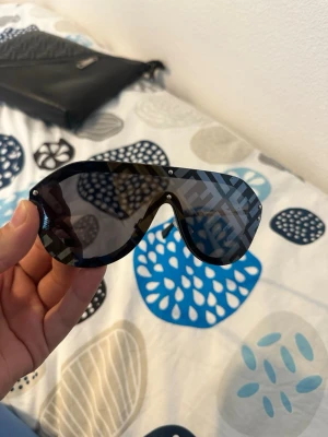 Svarta Fendi solglasögon med logomönster - Säljer ett par statement solglasögon från Fendi med svart båge och stora, rundade linser. Glasen har ett tydligt logomönster i grått och svart som ger en unik look. Snygga guldfärgade detaljer på skalmarna. Perfekta för dig som vill sticka ut.