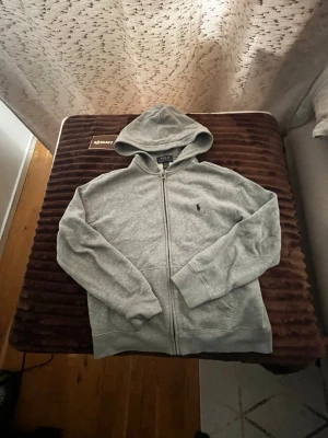 Grå zip hoodie från Polo Ralph Lauren - Snygg grå hoodie med dragkedja från Polo Ralph Lauren. Klassisk design med huva, två fickor framtill och den ikoniska lilla loggan broderad på bröstet. Tillverkad i mjukt bomullsmaterial som är perfekt för chill dagar eller när du vill ha en clean look.