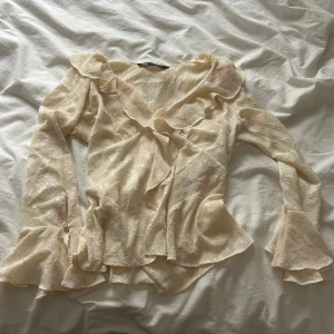 Krämvit volangblus med struktur - Romantisk krämvit blus med volangdetaljer längs halsringning och framtill. Långärmad modell med lätt transparent, strukturerat tyg och subtilt mönster. Perfekt för dig som gillar en feminin och trendig stil.