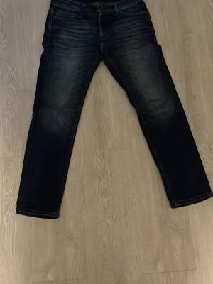 Mörkblå tapered jeans Mike - Säljer ett par mörkblå jeans med tapered fit, modell Mike. Jeansen har snygga slitningar och klassisk femficksdesign. Perfekta för dig som gillar en smalare passform vid anklarna och en modern look.