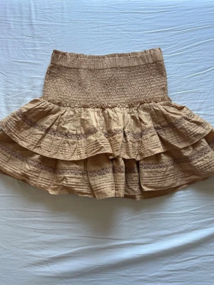 Beige volangkjol med spetsdetaljer - Supersöt kort volangkjol i beige med bred smock i midjan och fina spetsdetaljer på volangerna. 