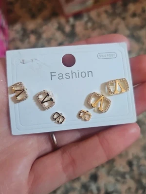 Guldiga örhängen med glittrande detaljer - Säljer ett set med tre par guldiga örhängen i olika storlekar. Alla har en fyrkantig form med ett V-inspirerat mönster och glittrande stenar runt kanterna. Perfekta för att mixa och matcha till olika looks.