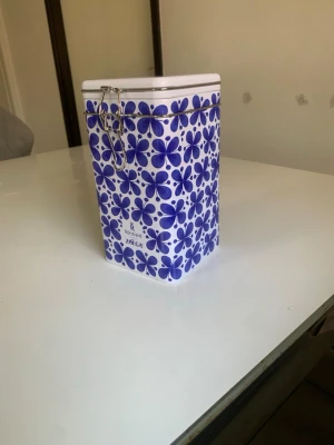 Blåvit plåtburk från Zoégas - Säljer en fyrkantig plåtburk från Zoégas med blått blommönster och vit bakgrund. Burken har ett metallspänne för förslutning och är perfekt för förvaring av kaffe eller te. Märket Zoégas syns tydligt på locket. Den är helt ny oanvänd.