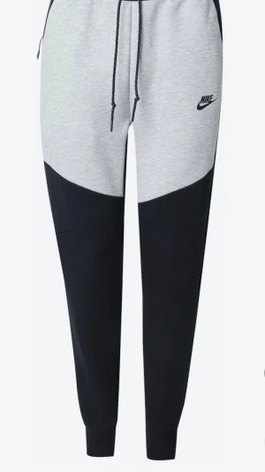 Nike mjukisbyxor i grått och svart - Snygga mjukisbyxor från Nike i grått och svart med dragsko i midjan och ribbade benslut. Byxorna har en modern passform och Nike-logga på låret. Perfekta för chill dagar eller träning.