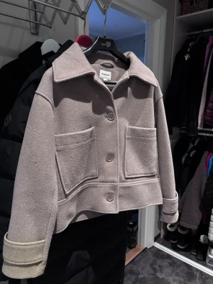 Vårkappa från Kappahl - Kort beige cardiganjacka från Kappahl i ullmix med stora fickor framtill och bred krage. Jackan har stora knappar och uppvikta ärmslut med knappdetalj. Perfekt för lager på lager och en clean look.