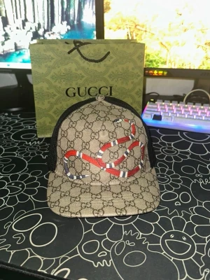 Gucci snake keps  - Snygg keps från Gucci med klassiskt GG-mönster i beige och brunt, dekorerad med en röd och vit orm på framsidan. Baksidan är i svart mesh och har justerbart spänne med grönt och rött band. Perfekt för dig som vill sticka ut med lyxig streetwear.
