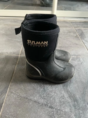 Svarta kängor från Sulman Footwear - Svarta kängor från Sulman Footwear med högt skaft i neopren och slitstark gummisula. Kängorna har vita detaljer på sidan och reflex baktill. Perfekta för kalla och blöta dagar.