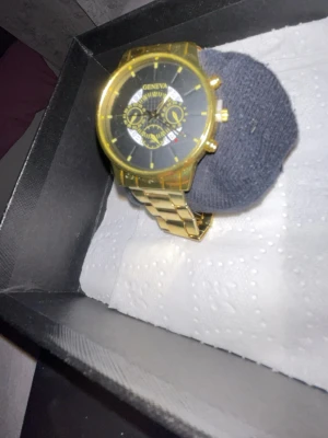 Guldig klocka från Geneva - Stilren klocka från Geneva med guldig metall-länk och svart urtavla. Klockan har flera dekorativa visare och detaljer i guld, vilket ger en lyxig känsla. Perfekt accessoar för dig som vill sticka ut med en elegant look.