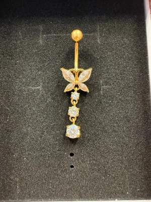 Guldfärgad navelpiercing fjäril - Snygg guldfärgad navelpiercing med en glittrande fjäril och tre hängande stenar. Piercingen passar perfekt för dig som vill ha något extra till din look.