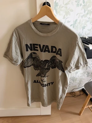 Beige t-shirt från Dsquared2 med tryck - Säljer en beige t-shirt från Dsquared2 med stort svart tryck framtill med texten 'NEVADA ALMIGHTY' och en örn. Klassisk rund halsringning och korta ärmar i storlek L. Inga hål eller defekter 