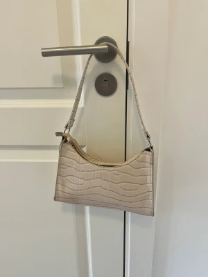 Beige axelremsväska i krokopräglat skinn - Snygg beige handväska med axelrem och krokodilpräglat mönster. Väskan har en stilren, avlång form och stängs med dragkedja upptill. Perfekt accessoar för att lyfta din outfit med en trendig touch.