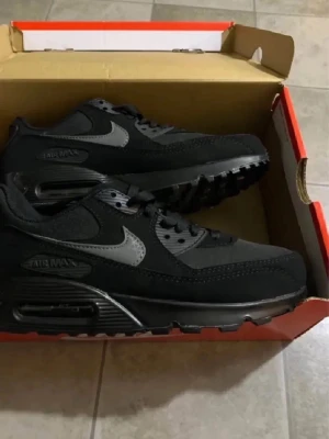 Nike Air Max 90 svarta sneakers - Svarta Nike Air Max 90 sneakers med grå swoosh och detaljer. Klassisk siluett med synlig Air Max-dämpning i sulan. Ovandel i mesh och läder för både stil och komfort. Perfekta för dig som gillar streetwear och sportig look.