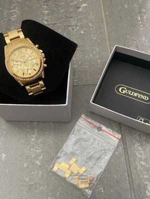 Guldig klocka från Guldfynd med Stenar - Snygg guldig klocka från O.N.E med glittrande stenar runt urtavlan och länkarmband i metall. Klockan har flera dekorativa urtavlor och kommer i originalask från Guldfynd. Extra länkar medföljer för justering av storlek. Snabb Leverans!