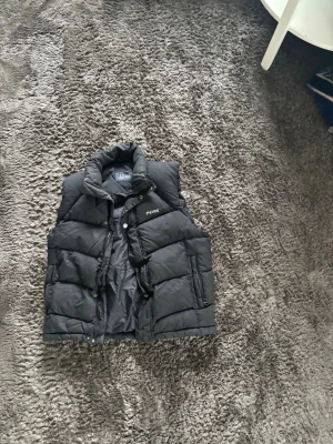 Svart dunväst från Peak Performance - Snygg svart dunväst från Peak Performance med hög krage och tryckknappar framtill. Västen har två stora fickor och diskret logga på bröstet. Perfekt för lager-på-lager och riktigt skön att bära under kyliga dagar.