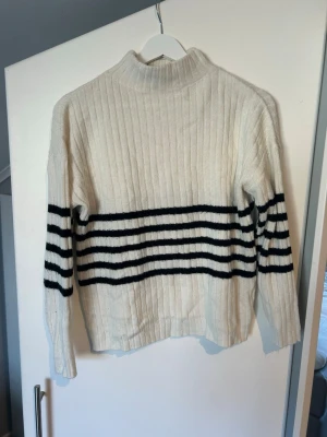 Vit stickad tröja med svarta ränder - Vit ribbstickad tröja med svarta horisontella ränder över bröstet och armarna. Tröjan har hög hals och långa ärmar, perfekt för kyligare dagar. Materialet ser mjukt och mysigt ut, och modellen är något boxig för en avslappnad look.