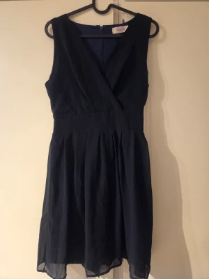 Mörkblå omlottklänning med knyt - Snygg mörkblå klänning med omlott framtill och knytband i midjan. Klänningen är ärmlös och har en lätt, luftig känsla med vippig kjol. Perfekt för dig som gillar stilrena och klassiska plagg med en feminin touch.