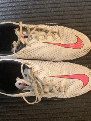 Vita Nike fotbollsskor med rosa swoosh - Säljer ett par vita Nike fotbollsskor med rosa swoosh på sidan och sulan. Skorna har snörning, låg profil och mönstrad ovandel för bättre. Perfekta för dig som vill sticka ut på matchen och som vill ha bra bollkontroll inomhus 