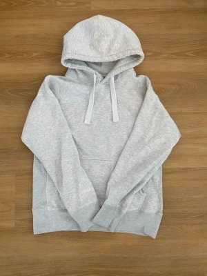 Ljusgrå hoodie från Russemerch  - Ljusgrå hoodie från Russedress med klassisk huva och dragsnören. Hoodien har en stor magficka och ribbade muddar vid ärmslut och nederkant. Tillverkad i mjukt sweatshirtmaterial, perfekt för en avslappnad stil.