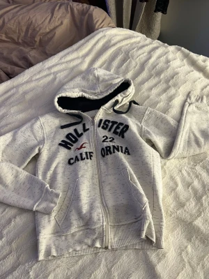 Vit hoodie från Hollister med dragkedja - Vit hoodie från Hollister med svart text och logga på bröstet, dragkedja framtill och två fickor. Mjuk insida och svart fodrad huva med snören. Perfekt för en avslappnad och cool stil.