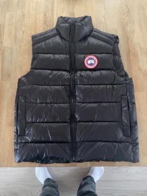 Svart dunväst från Canada Goose - Säljer en svart dunväst från Canada Goose med klassisk logotyp på bröstet. Västen har hög krage, raka fickor och quiltad design. Perfekt för lager-på-lager och håller dig varm under kyliga dagar. Stängs med dragkedja framtill. Kan diskuteras om priset.