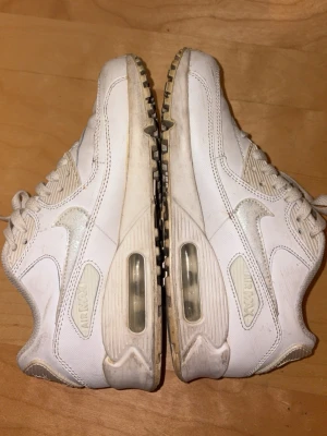 Nike Air Max 90 vita sneakers - Klassiska vita Nike Air Max 90 sneakers med synlig luftdämpning i sulan och ikonisk swoosh på sidan. Skorna har snörning och är tillverkade i en mix av läder och syntetmaterial. Perfekta för dig som gillar streetwear och sportig stil.