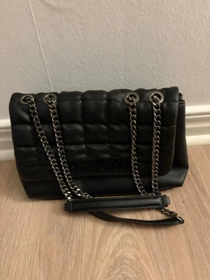 Svart quiltad axelväska med kedjerem - Snygg svart axelväska i quiltad design med dubbla silverfärgade kedjeremmar och vadderad yta. Väskan har två stora fack och innerficka med dragkedja. Perfekt för dig som gillar trendiga accessoarer med lite edge.