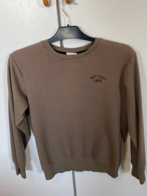 Beige sweatshirt från Kappahl - Säljer en beige sweatshirt från Kappahl med texten 'West Coast 1978' broderad på bröstet. Tröjan har rund halsringning, ribbade muddar och ett diskret rutmönster framtill. Perfekt för en avslappnad stil.