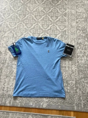 Blå t-shirt från Polo Ralph Lauren - Snygg blå t-shirt från Polo Ralph Lauren med klassisk broderad logga på bröstet. T-shirten har rund halsringning och korta ärmar. Tillverkad i mjuk bomull som känns skön mot huden. Perfekt för en avslappnad och stilren look.