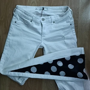 Vita jeans med svart prickigt tyg - Unika vita jeans från Abercrombie & fitch med svart tyg och stora vita prickar längs ena benet. Klassisk femficksmodell med knappgylf och raka ben. Snygg kontrastdetalj som sticker ut och ger en cool retrovibe. Perfekta för dig som vill ha något annorlunda i garderoben. Stdda bootcut. Sorlek 27
