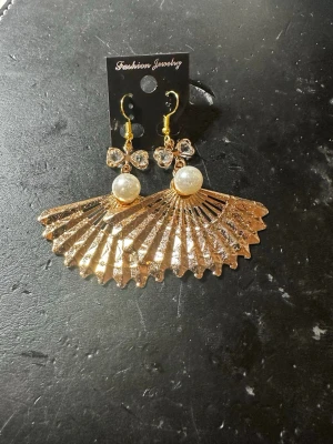 Guldiga solfjäderörhängen med pärlor - Unika örhängen i guldmetall med solfjäderformad design, dekorerade med vita pärlor och gnistrande stenar. Perfekta för dig som vill sticka ut med accessoarer som har både elegans och attityd. Krokar i guldfärg för enkel användning.