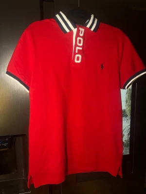 Röd pikétröja från Polo Ralph Lauren - Röd pikétröja från Polo Ralph Lauren med vit och marinblå krage och ärmslut. Stort 'POLO'-tryck vid knäppningen och klassisk logga broderad på bröstet. Perfekt för en sportig och stilren look.
