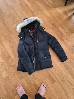 Svart dunjacka från Moose Knuckles - Snygg svart dunjacka från Moose Knuckles med avtagbar huva och vit fuskpäls. Jackan har flera dragkedjeförsedda fickor, hög krage och detaljer i silver. Perfekt för kalla dagar och riktigt varm tack vare dunfyllningen.