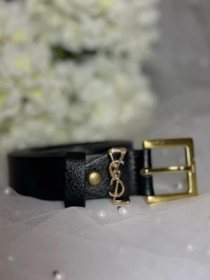 Svart YSL bälte med gulddetaljer - Stilrent svart bälte från Yves Saint Laurent i läder med elegant guldfärgat spänne och ikonisk YSL-logga i metall. Perfekt accessoar för att lyfta din outfit med en lyxig touch.