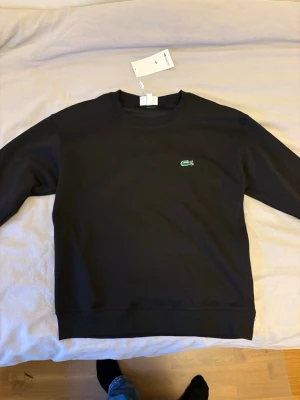 Svart Lacoste sweatshirt - Snygg svart sweatshirt från Lacoste med klassisk rund hals och det ikoniska gröna krokodilmärket broderat på bröstet. Tröjan har långa ärmar och ribbade muddar vid ärmslut och nederkant. Tillverkad i mjukt bomullsmaterial för skön komfort. Helt ny, aldrig använd