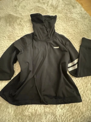 Svart hoodie från Stronger - Svart hoodie från Stronger med hög krage och dragsko. Tröjan har en sportig look med vita ränder på ena ärmen och logga på bröstet. Tillverkad i ett mjukt och stretchigt material, perfekt för en aktiv vardag.