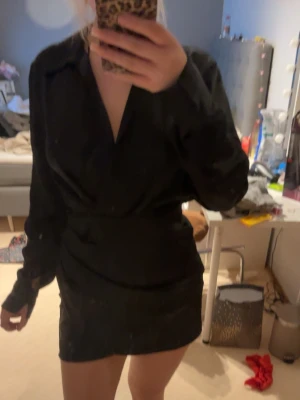 Svart omlottklänning med v-ringning - Snygg svart kortklänning med omlott och v-ringad hals. Klänningen har långa ärmar och markerad midja för en stilren siluett. Perfekt för dig som gillar en enkel men elegant look.