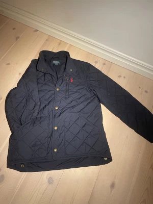  quiltad jacka från Polo Ralph Lauren - Mörkblå quiltad jacka från Polo Ralph Lauren med guldiga knappar, hög krage och dragkedja vid bröstfickan. Jackan har röd broderad logga på bröstet och långärmad passform. Materialet är troligtvis polyester och jackan har en klassisk, stilren look.