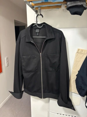 Svart overshirt med dragkedja - Stilren svart overshirt från COS med rak passform, två stora bröstfickor och silverfärgad dragkedja framtill. Overshirten har klassisk krage och långa ärmar med breda manschetter. Perfekt för lager-på-lager och enkel att matcha.