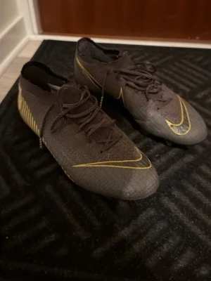 Nike Mercurial svarta/gula fotbollsskor - Säljer ett par Nike Mercurial fotbollsskor i svart med gula detaljer och snörning. Skorna har en strömlinjeformad design, mönstrad ovandel och fasta dobbar för bra grepp på planen. Tillverkade i syntetmaterial för lätt känsla och bra bollkontroll.